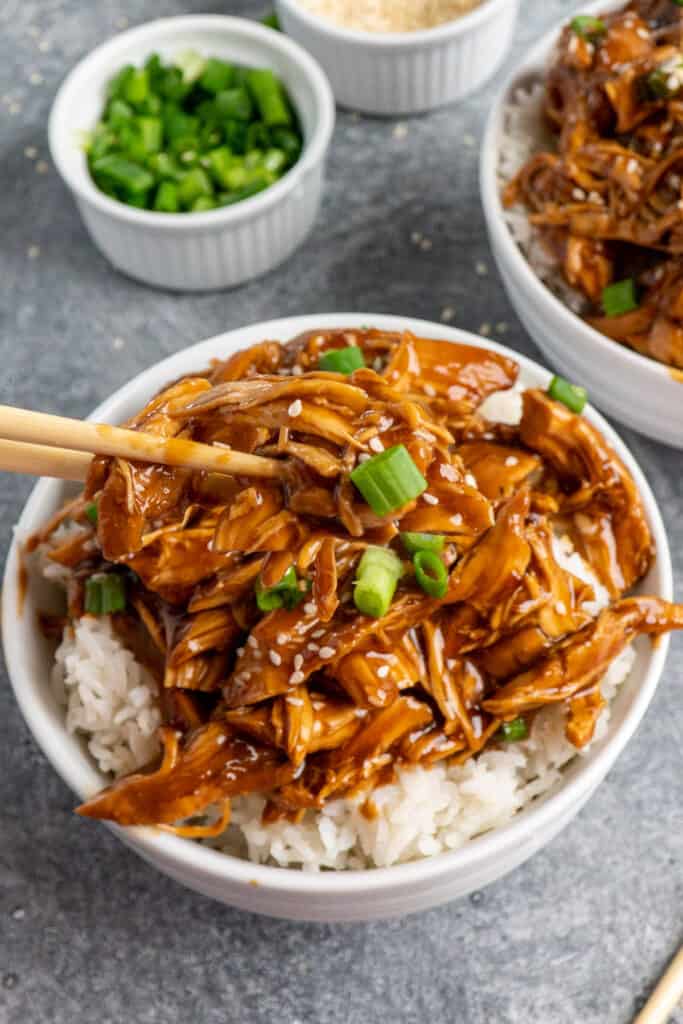 Close up of chop sticks holding shredded slow cooker honey teriyaki chicken in a white bowl over rice.