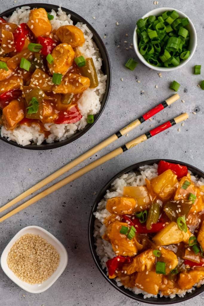 Two bowls of sweet and sour chicken over rice garnished with sesame seeds and green onions.