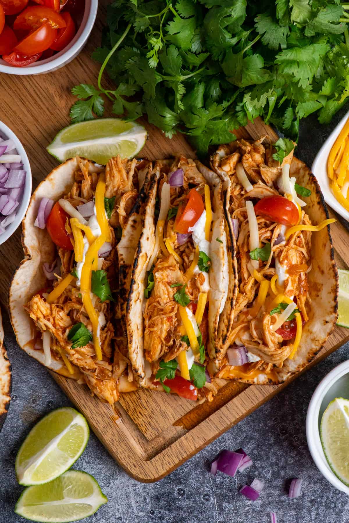 Three shredded chicken crockpot tacos on a wood cutting board with fresh toppings and a bunch of cilantro and bowl of tomatoes on the side.