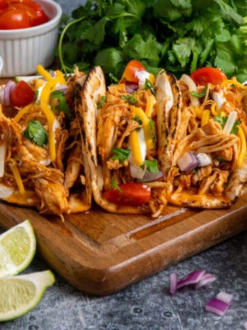 Close up of 3 slow cooker shredded chicken tacos on a wood cutting board.