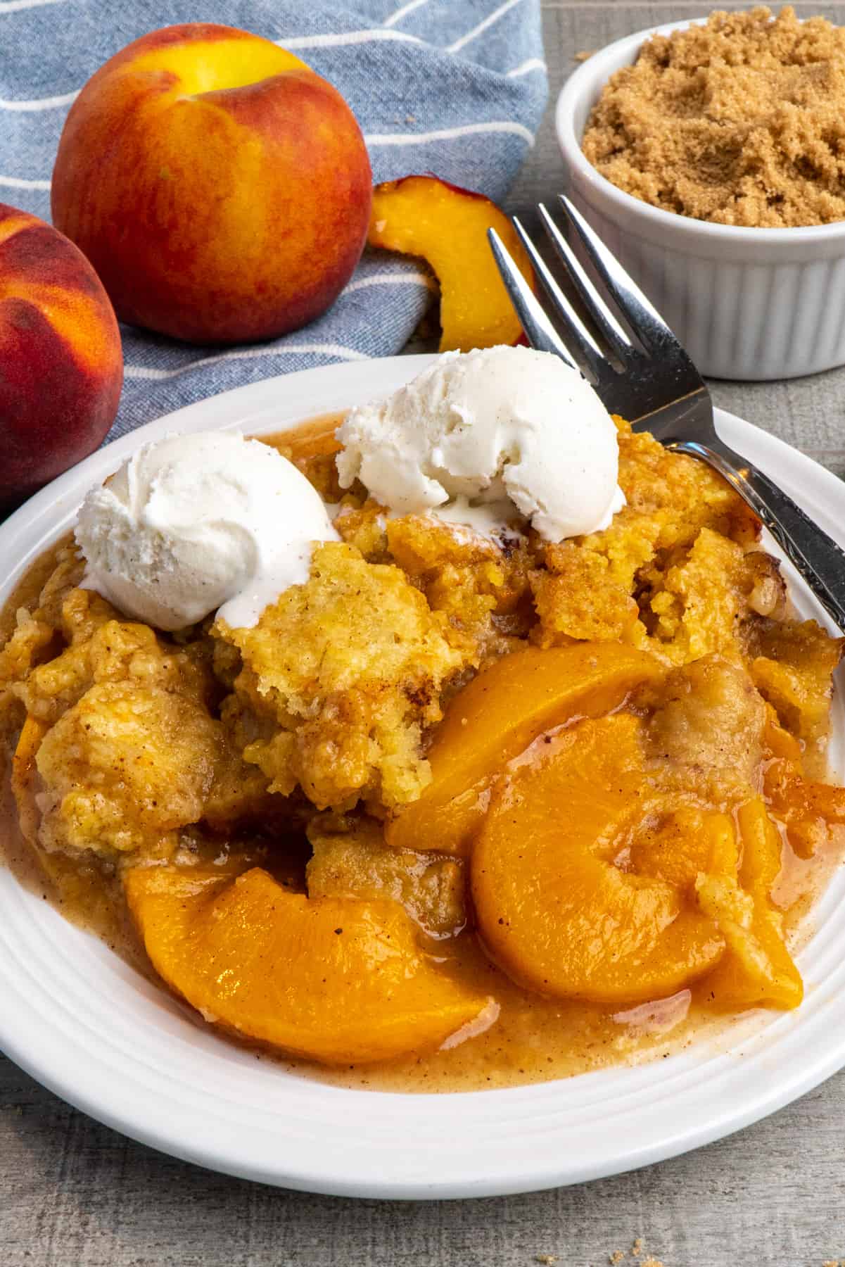 Crock-Pot peach cobbler on a white plate topped with ice cream and whole peaches in the background.