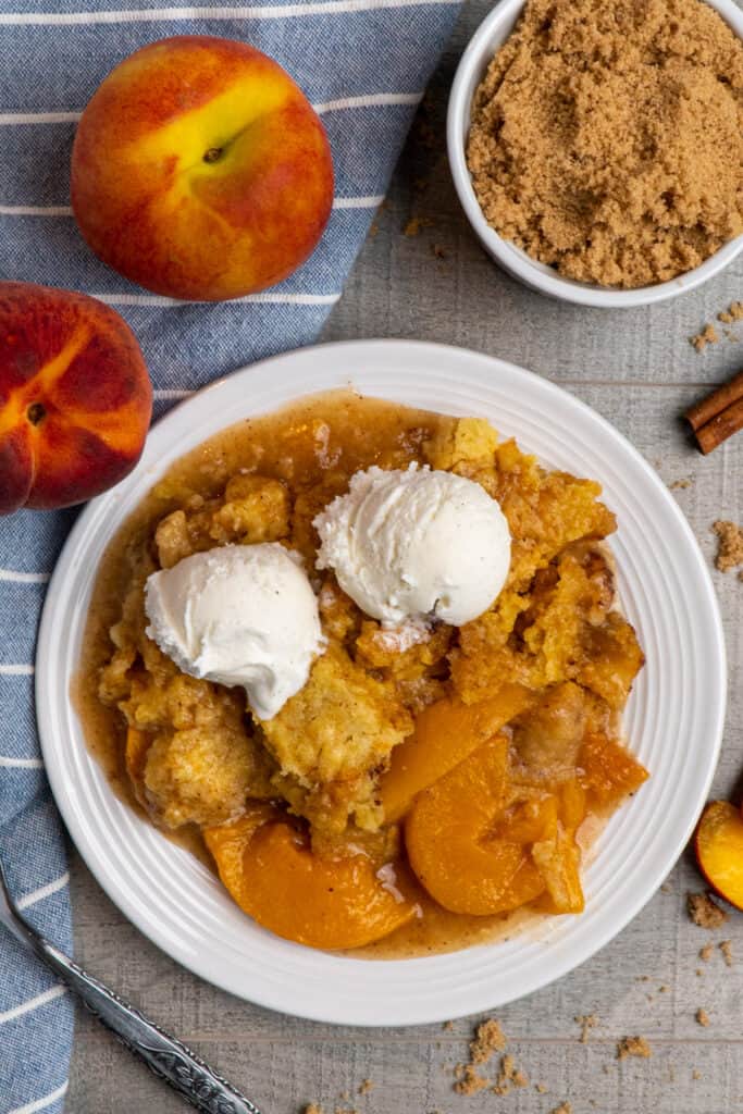 Over head look at slow cooker peach cobbler on a white plate topped with ice cream and fresh peaches in the background.