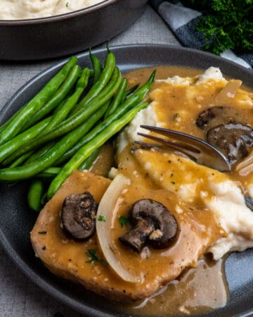 Crock-Pot smothered pork chops on a grey plate with green bean and mashed potatoes with gravy.