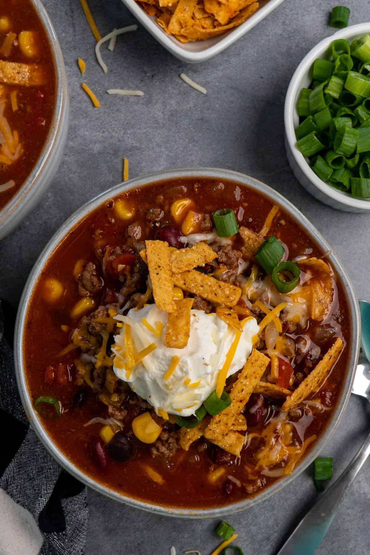 Bowl of slow cooker taco soup topped with sour cream, green onions, cheese and tortilla strips on a table with small bowls of ingredients around it.