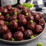 Close up of a Crockpot meatballl with grape jelly and chili sauce on a grey plate and garnished with green onions.