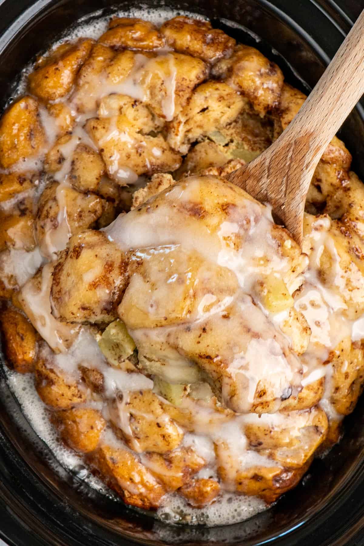 Apple cinnamon rolls being held with a wooden spoon over a slow cooker.