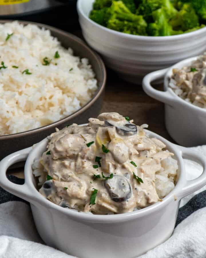 Slow cooker artichoke mushroom chicken in a white bowl over rice and broccoli in the background.