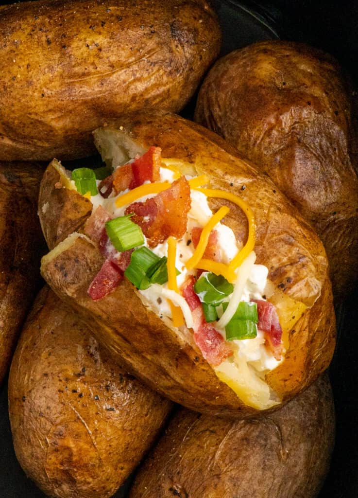 Close up of a loaded baked potato in a slow cooker on top of other potatoes.