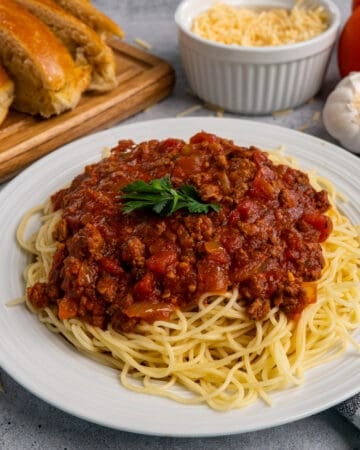 Close up of slow cooker meat sauce over spaghetti noodles on a white plate with bread in the background.