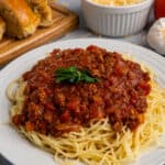 Close up of slow cooker meat sauce over spaghetti noodles on a white plate with bread in the background.