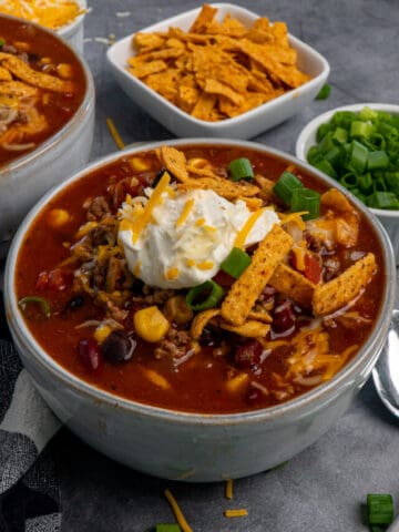 Close up of Crock-Pot taco soup in a grey bowl and topped with sour cream, green onions, cheese and tortilla strips.