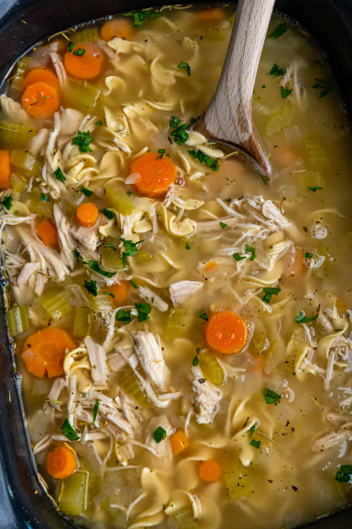 Overhead look at chicken noodle soup in a slow cooker with a wooden spoon in it.
