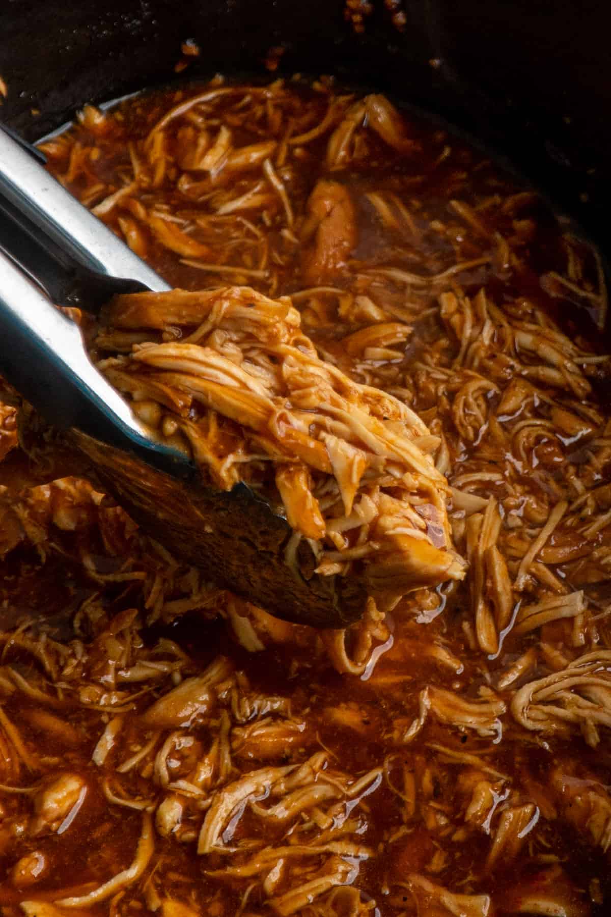 Close up of Dr. Pepper chicken being held by tongues over a slow cooker of shredded chicken.
