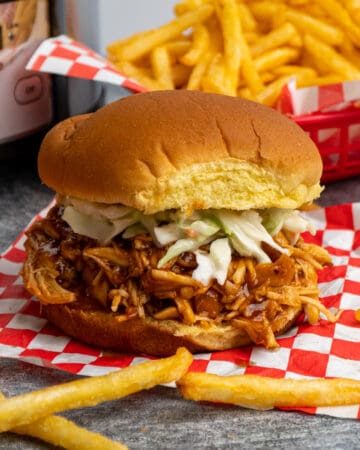 Close up of Crock Pot Dr. Pepper Chicken on a potato bun with fries in the background.
