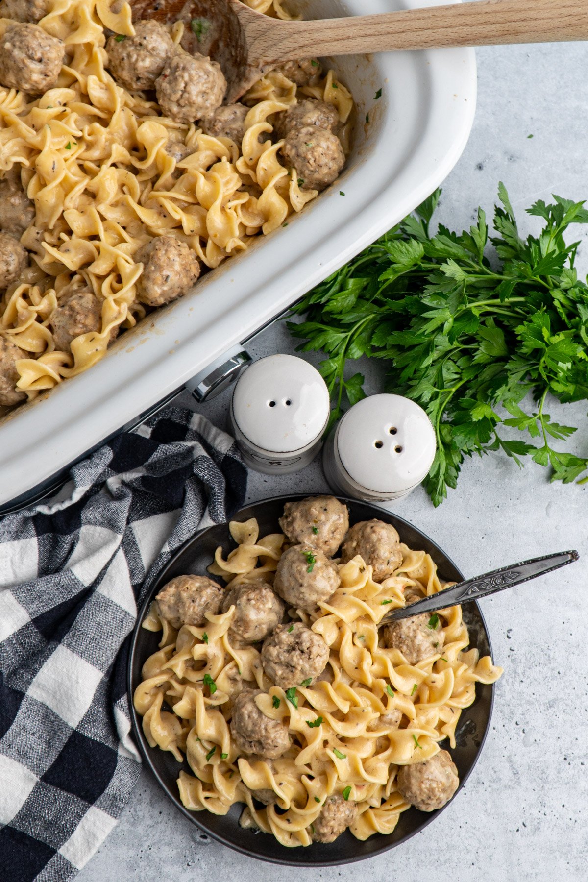 Swedish meatballs on a plate and in a slow cooker on the table with a bunch of parsley to the side.