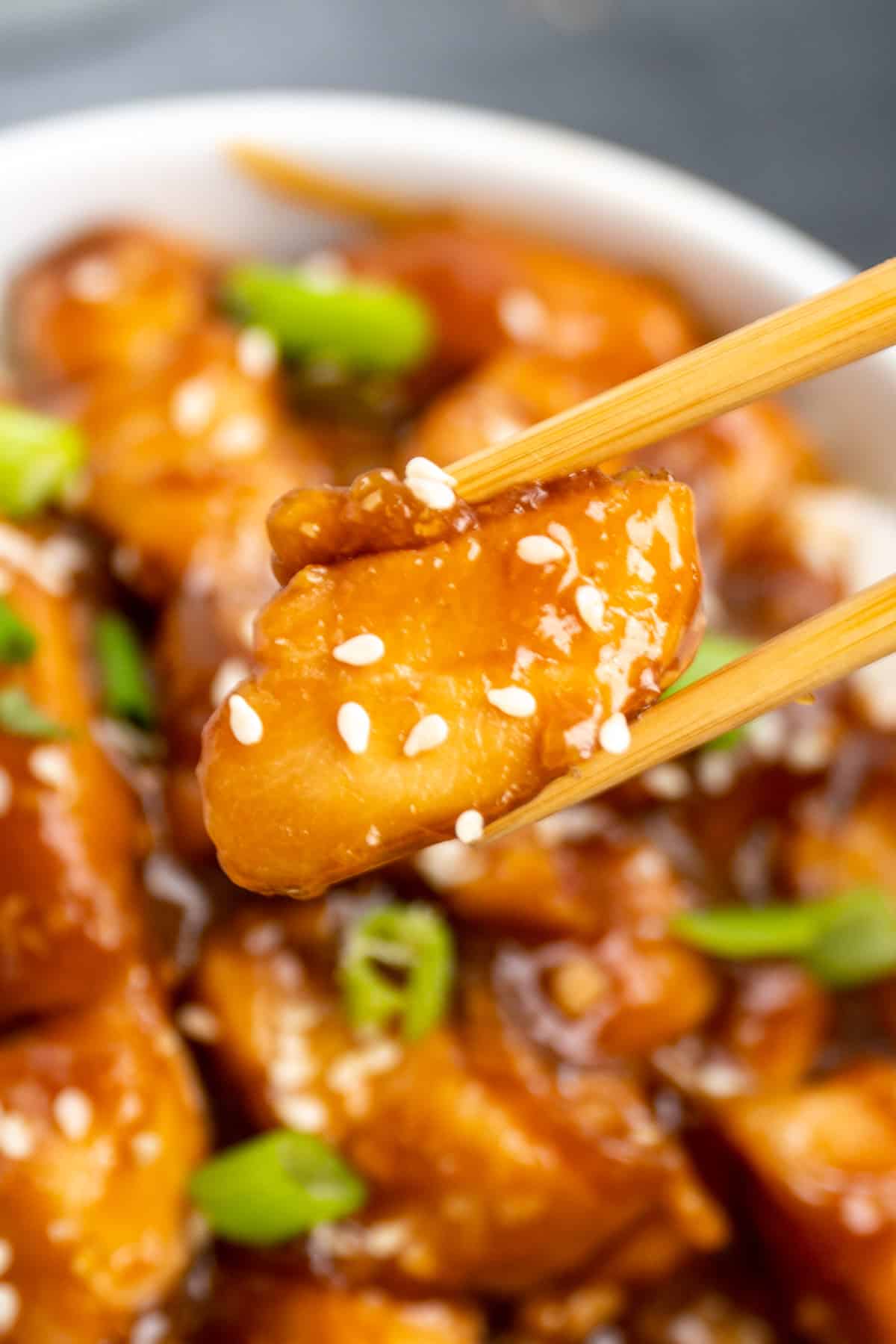 Close up of chopsticks holding general Tso's chicken over a bowl of chicken.