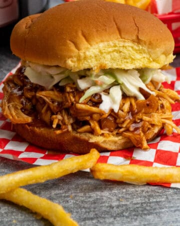 Close up of Crock Pot Dr. Pepper Chicken on a potato bun with fries in the background.
