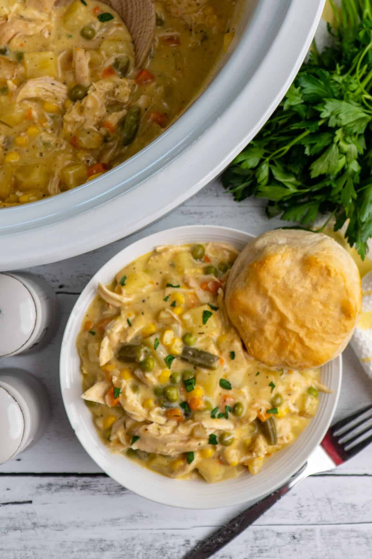 Overhead look at chicken pot pie in a white bowl and slow cooker with parsley in the background.