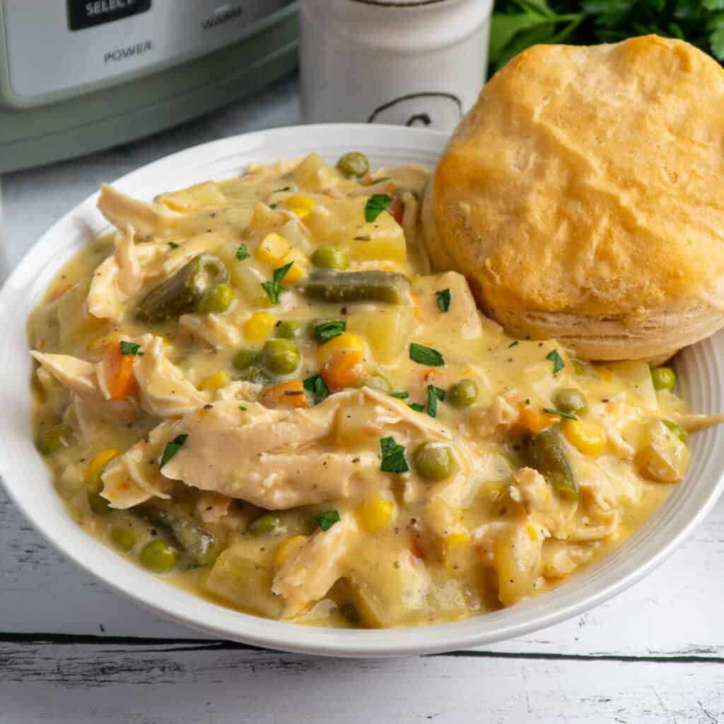 Crock Pot chicken pot pie in a white bowl with a biscuit in it and a slow cooker in the background.