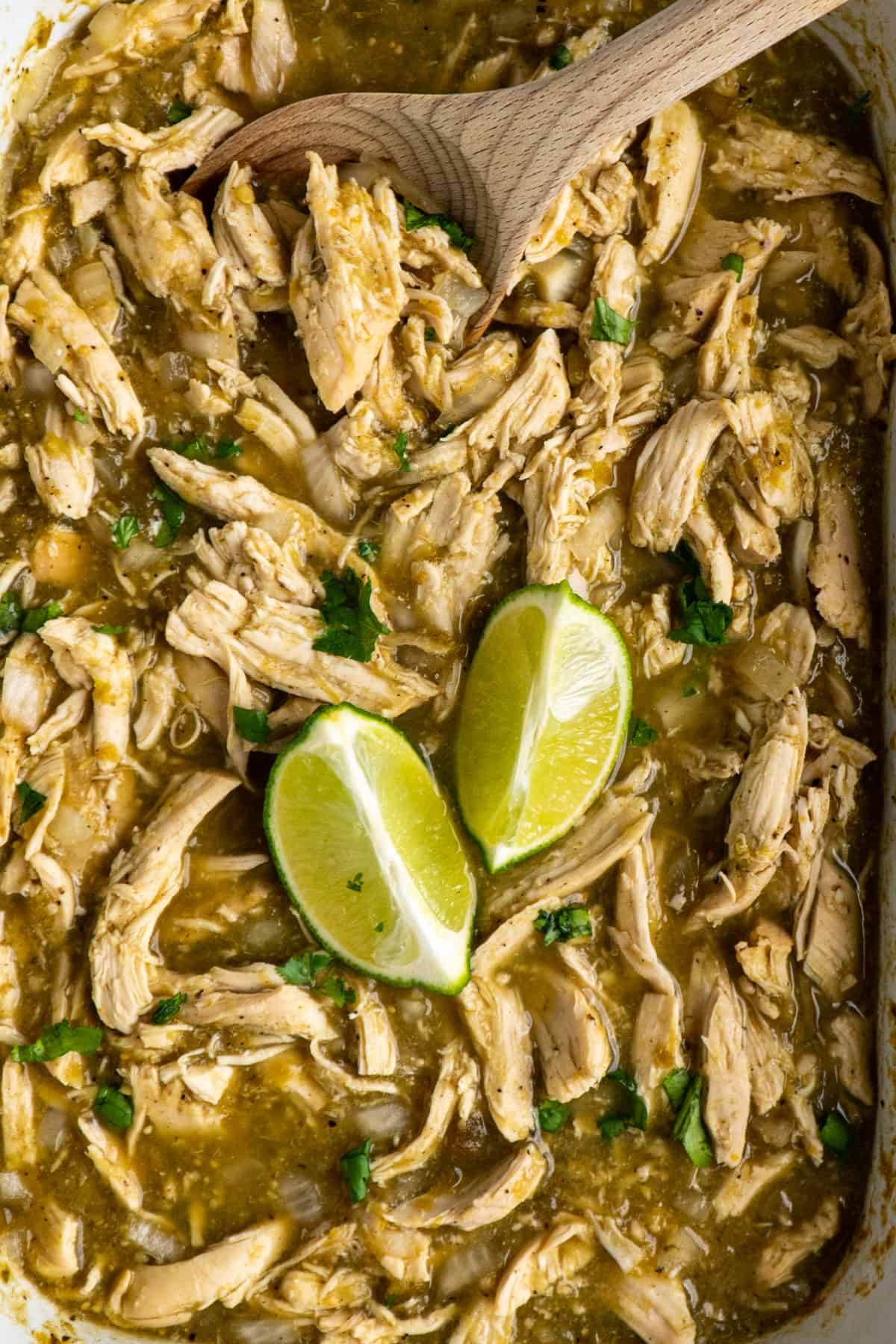 Salsa verde chicken in a slow cooker with a wooden spoon and two slices of lime.