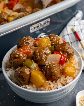 Close up of Crock-Pot sweet and sour meatballs over rice in a white bowl and garnished with sesame seeds.