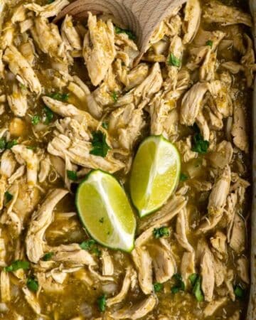 Salsa verde chicken in a slow cooker with a wooden spoon and two slices of lime.