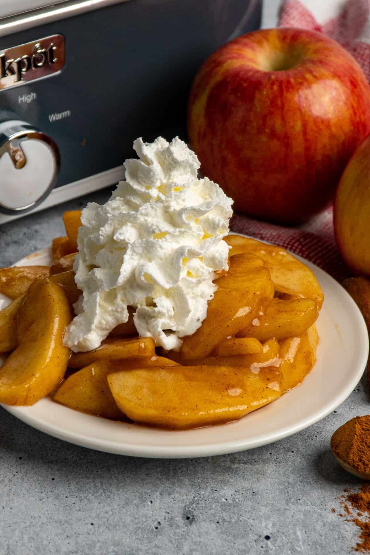 Close up of Cracker Barrel fried apples on a white plate with whipped cream on top and a slow cooker in the background.