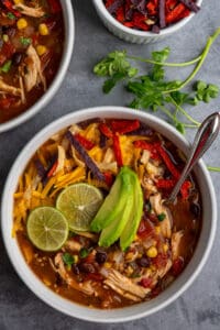 Crock Pot chicken tortilla soup in a bowl with limes and sliced avocado on top and tortilla strips in the background.