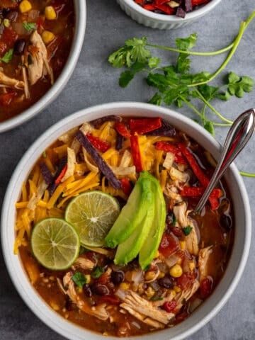 Crock Pot chicken tortilla soup in a bowl with limes and sliced avocado on top and tortilla strips in the background.