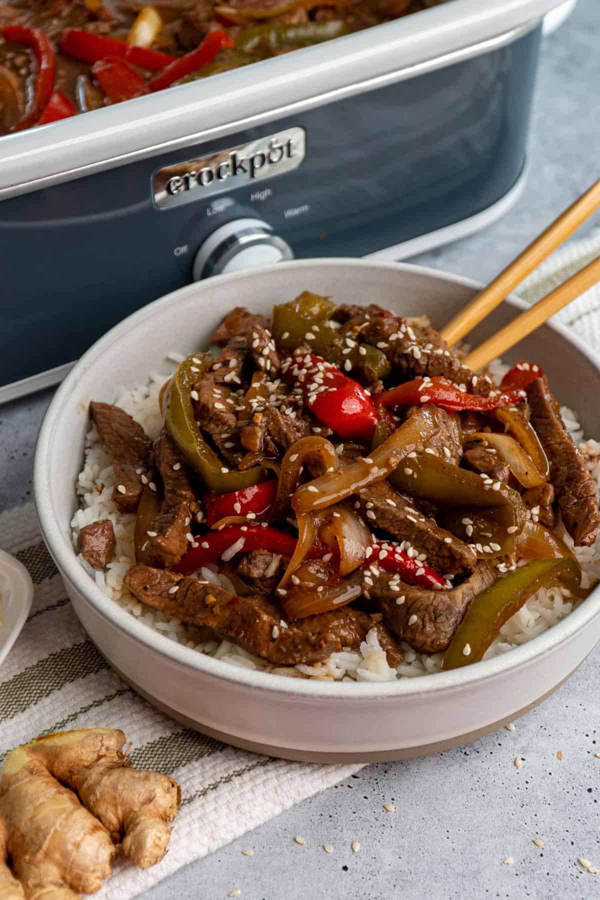 Beef and peppers over rice in a bowl with a slow cooker in the background.