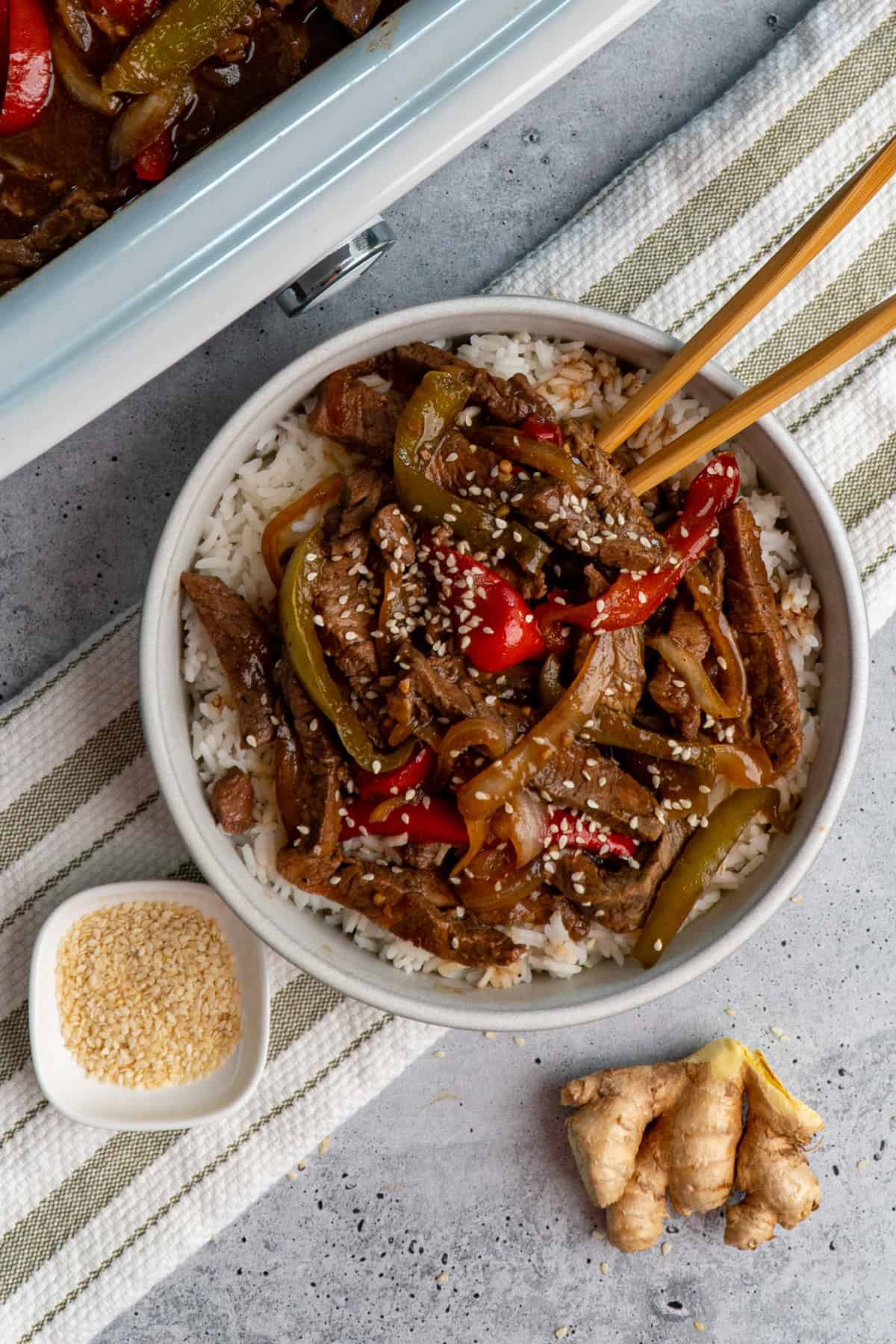 Overhead look at crock pot pepper steak over white rice in a bowl with ginger and sesame seeds on the side.