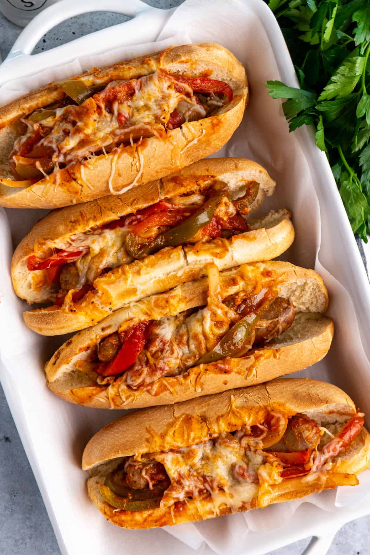Overhead look at crock pot sausage and peppers in hoagies covered in cheese inside a baking dish.
