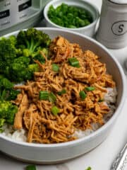 Close up of slow cooker honey garlic chicken in a bowl over rice with broccoli and a crock pot in the back ground.