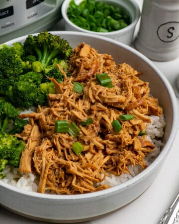 Close up of slow cooker honey garlic chicken in a bowl over rice with broccoli and a crock pot in the back ground.
