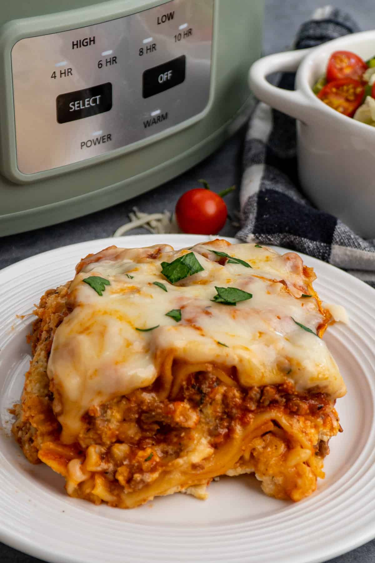 Close up of lasagna on a plate with a Crock-Pot and salad in the background.