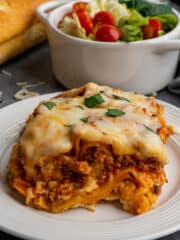 Slow cooker lasagna on a white plate with garlic bread and salad in the background.