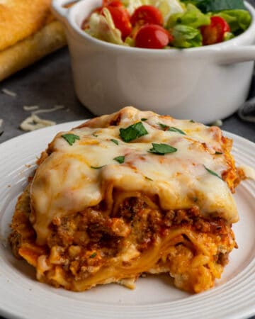 Slow cooker lasagna on a white plate with garlic bread and salad in the background.