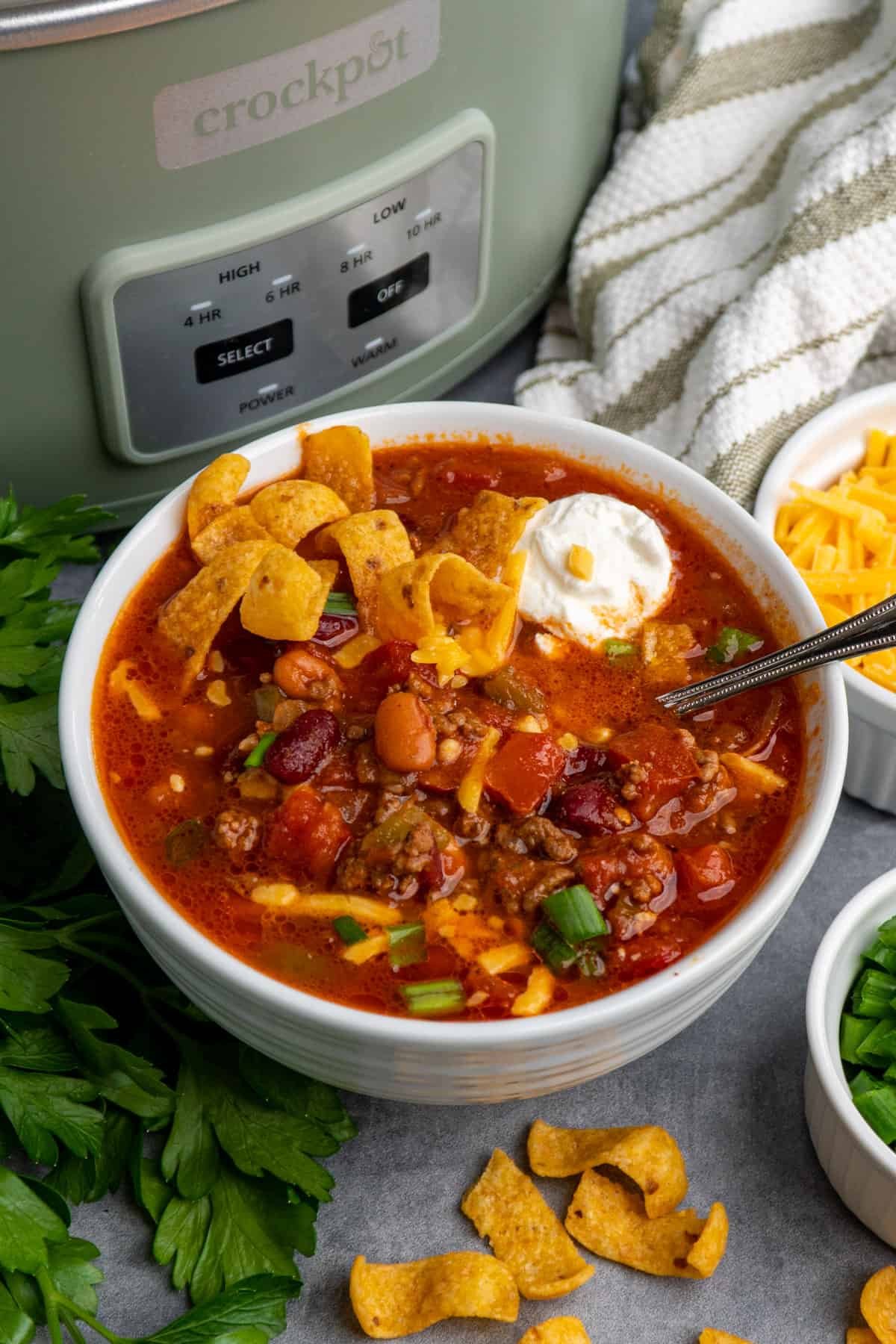The best chili in a white bowl topped with sour cream and cheese with a slow cooker in the background.