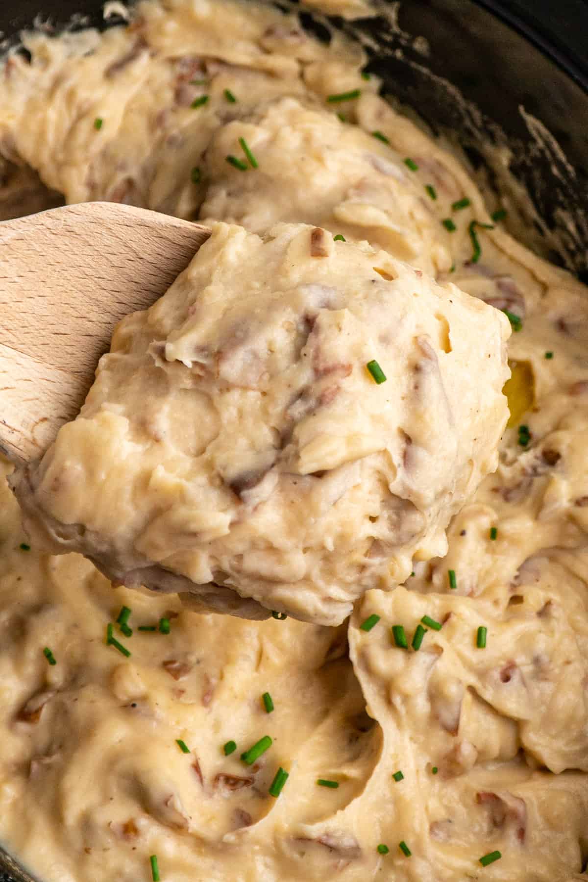 Close-up of creamy garlic mashed red potatoes with the skins on a wooden spoon over a slow cooker.