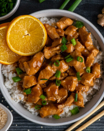 Slow cooker orange chicken over a bowl of rice garnished with orages and green onions.