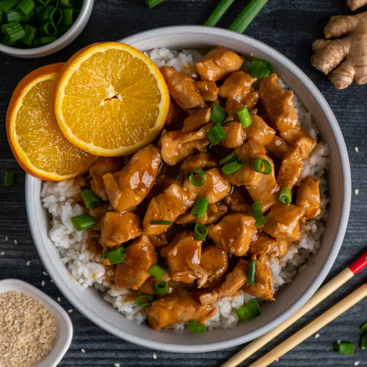 Slow cooker orange chicken over a bowl of rice garnished with orages and green onions.