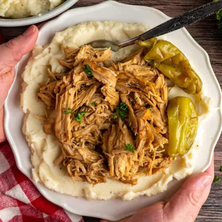 Hands holding a plate with Crock Pot Mississippi chicken on mashed potatoes.