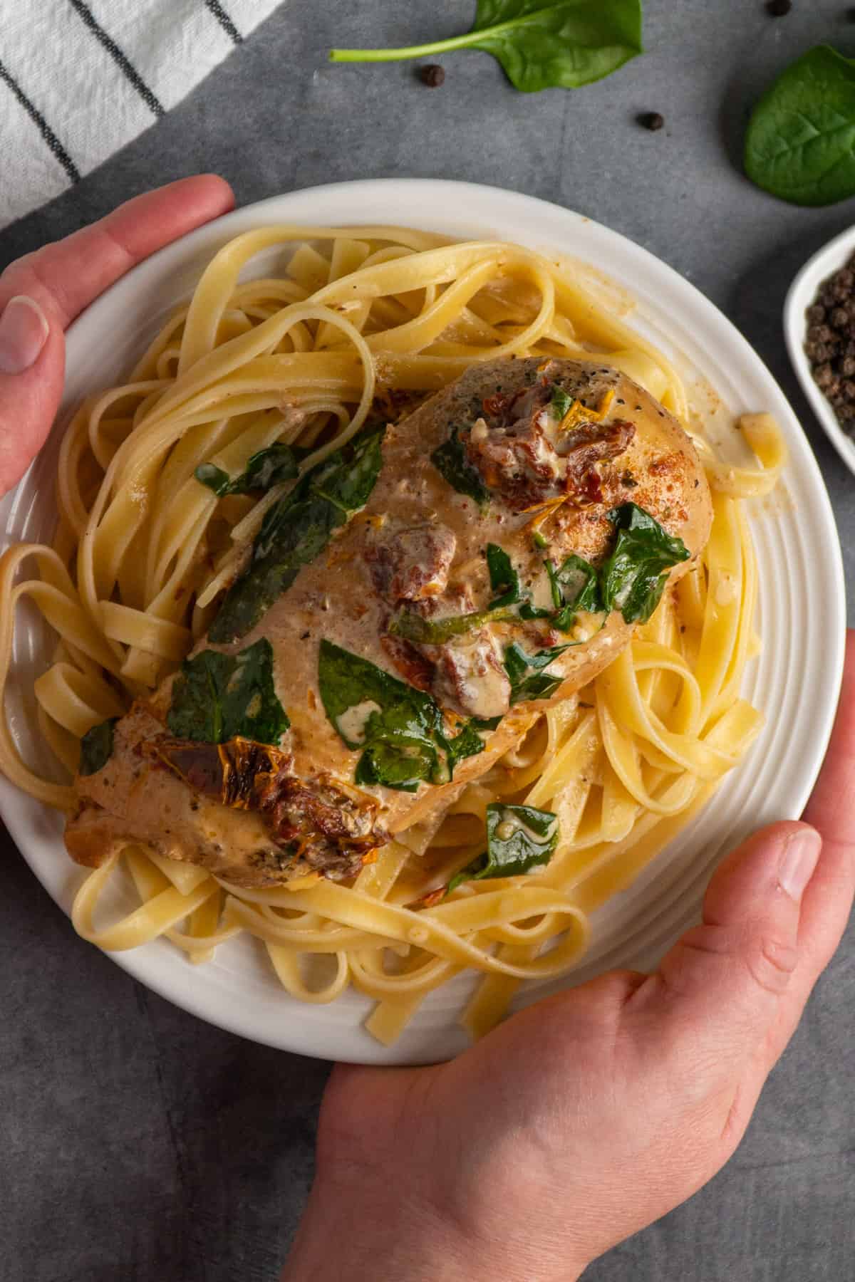 Two hands holding tuscan chicken on a plate of pasta.