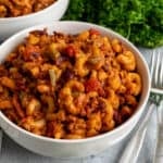 Crock pot chili mac in a white bowl with a fork on the side and a handful of parsley in the background with another bowl.