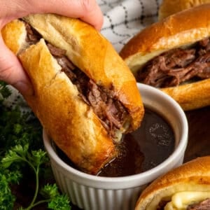 A hand dipping a crock pot French dip sandwich into au jus sauce.