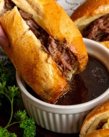 A hand dipping a crock pot French dip sandwich into au jus sauce.