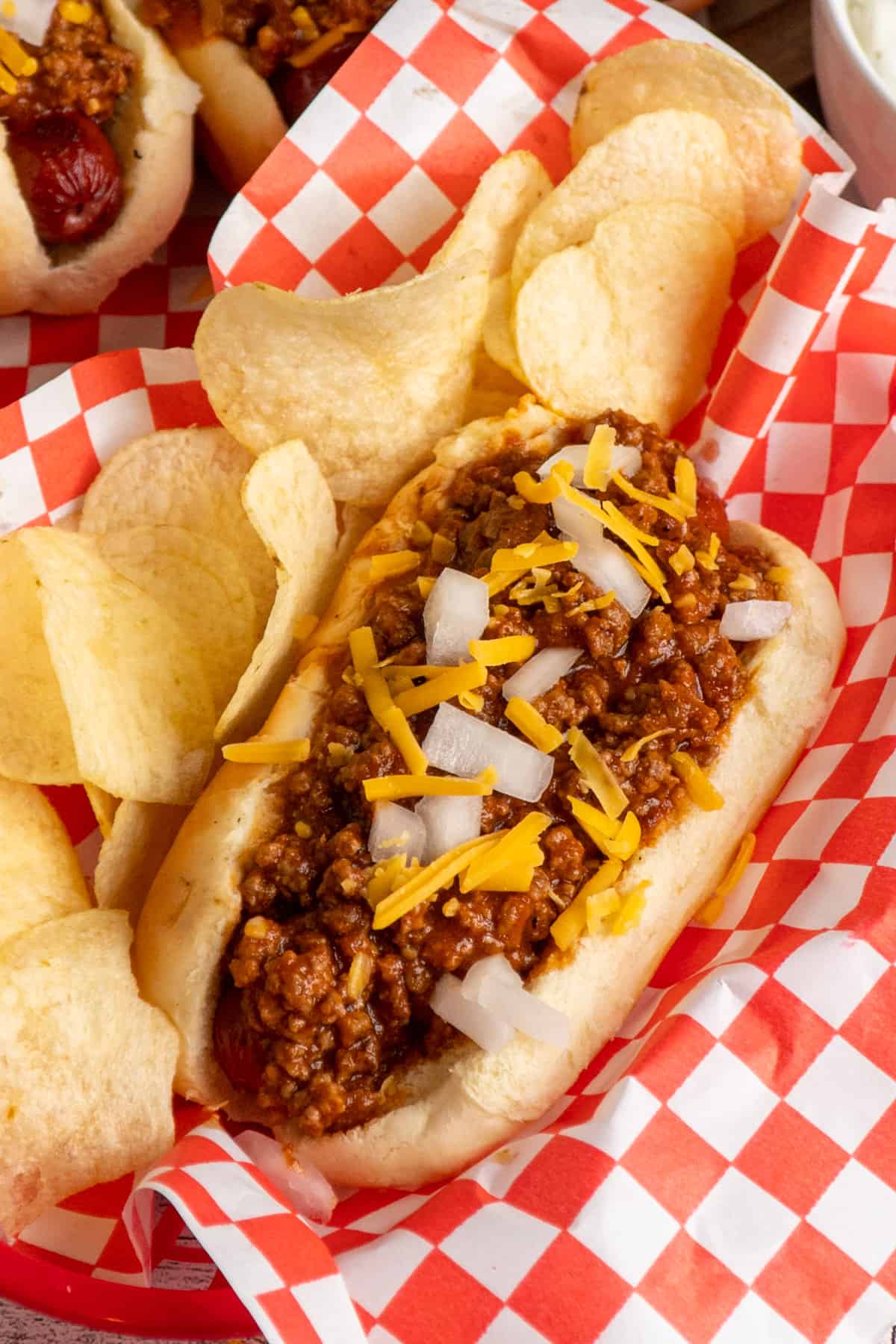 A hot dog in a basket topped with cheese and onions and served with chips.