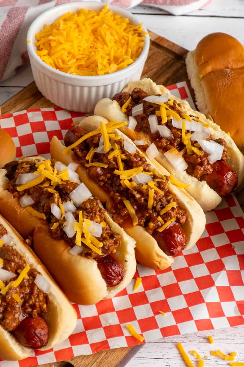 Four hot dogs on a wood cutting board and topped with chili and onions.