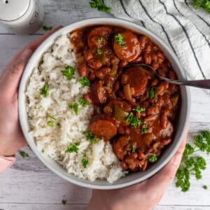 Hands holding a bowl of red beans and rice garnished with parsley.
