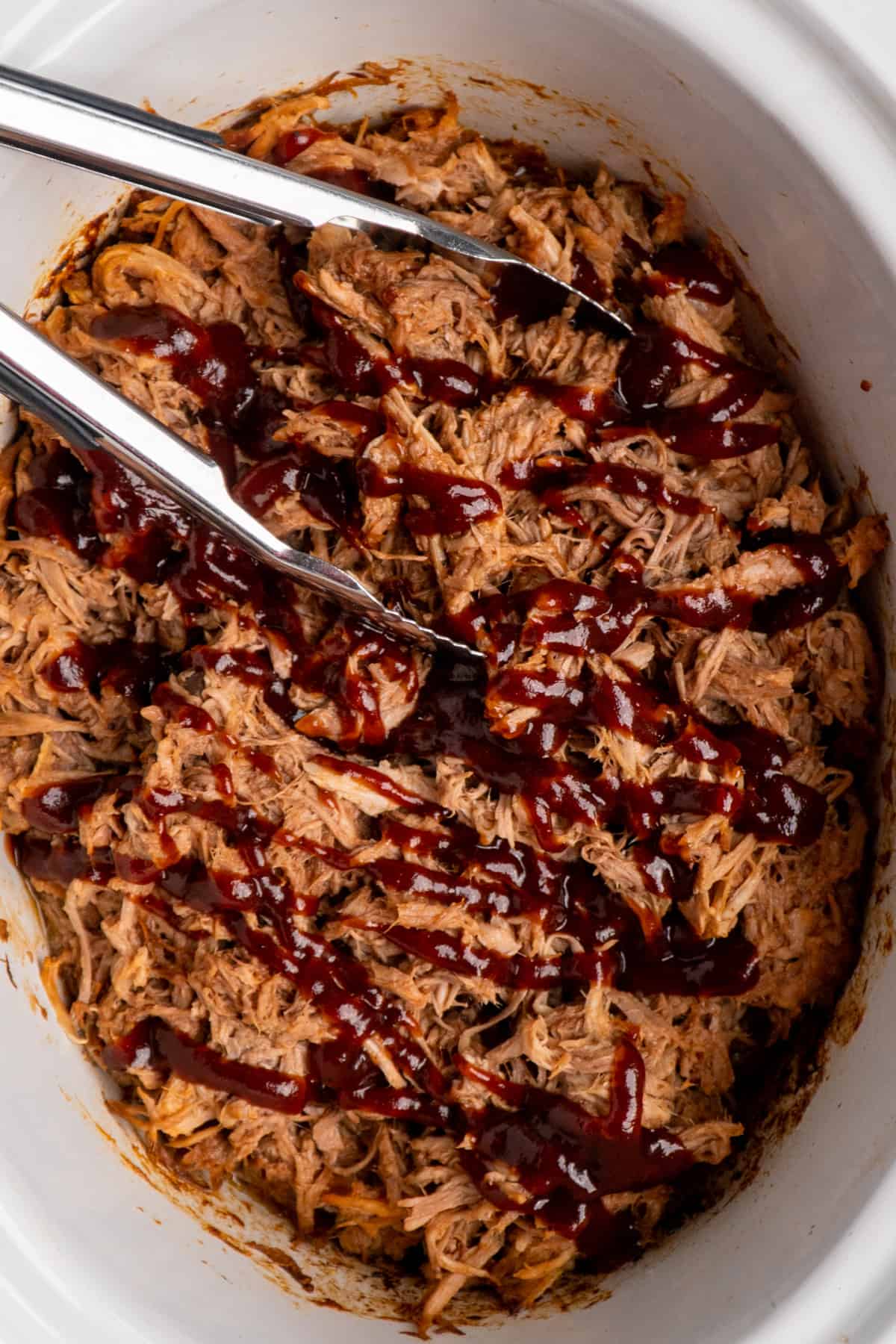 Shredded bbq pork with tongs in a white slow cooker with drizzled barbecue sauce.